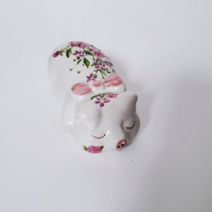May include: A white ceramic pig figurine with pink flowers and a pink bow. The pig is lying down with its eyes closed.