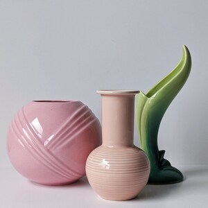 Op de afbeelding: Drie keramische vazen in roze en groen. De vaas links is een ronde, roze vaas met een geribbeld ontwerp. De vaas in het midden is een ronde, roze vaas met een geribbeld ontwerp. De vaas rechts is een groene vaas met een gebogen, bladvormig ontwerp.