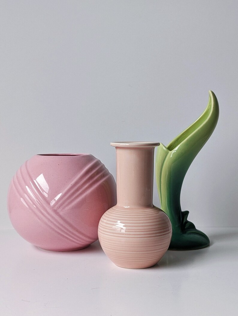 Op de afbeelding: Drie keramische vazen in roze en groen. De vaas links is een ronde, roze vaas met een geribbeld ontwerp. De vaas in het midden is een ronde, roze vaas met een geribbeld ontwerp. De vaas rechts is een groene vaas met een gebogen, bladvormig ontwerp.