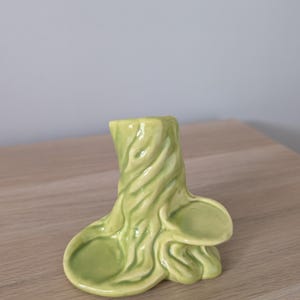 May include: A light green ceramic vase in the shape of a tree trunk with a textured, swirling design. The vase has a flared base with two rounded extensions and a small opening at the top.
