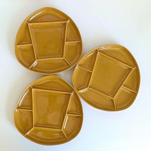 May include: Three mustard-yellow ceramic serving dishes. Each dish has a central square compartment and six surrounding compartments. The dishes have a unique, organic shape. The background is white.
