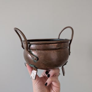 May include: A small, antique copper cauldron with two arched handles and three small feet. The cauldron has a rounded body and a dark interior. The copper has a rich, aged patina, with some areas showing a greenish hue. A decorative piece.