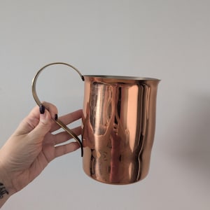 May include: A copper-coloured mug with a brass handle. The mug has a cylindrical body and a slightly flared rim. The handle is a simple, curved design. The mug is reflective, showing the surrounding environment. The mug is likely used for drinking beverages.