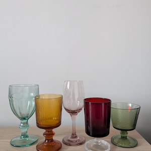 May include: Five colorful wine glasses in shades of green, amber, pink, burgundy, and green. The glasses have different shapes and textures, with stems and bowls. They are arranged on a light wooden surface.