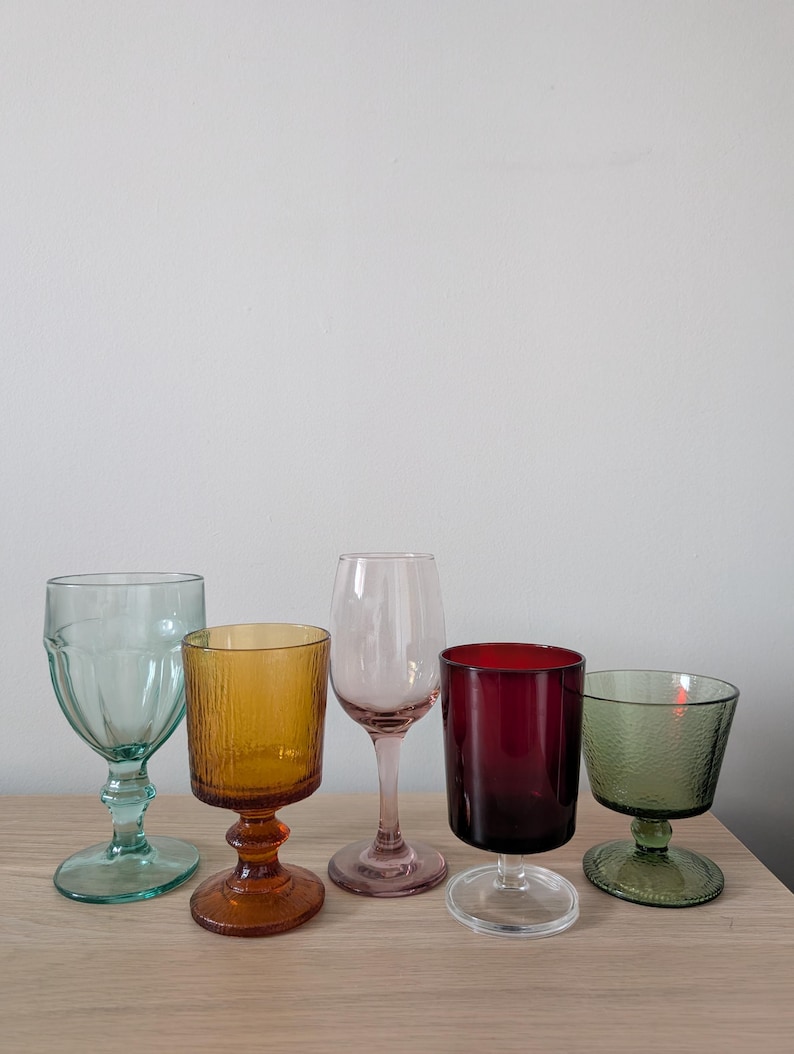 May include: Five colorful wine glasses in shades of green, amber, pink, burgundy, and green. The glasses have different shapes and textures, with stems and bowls. They are arranged on a light wooden surface.
