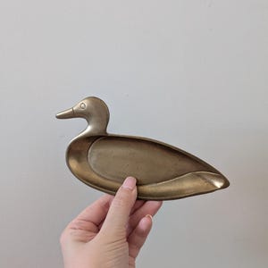 May include: A brass duck-shaped trinket dish. The dish is a golden colour and has a detailed duck head and body. The dish is held by a hand against a plain white background. The dish is designed to hold small items.