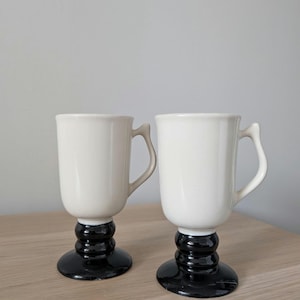 May include: Two white ceramic mugs with black pedestal bases and handles. The mugs have a classic design, with a cylindrical body and a curved handle. The black bases add a touch of contrast and elegance to the overall look.