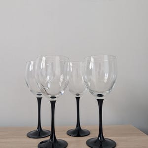 Op de afbeelding: Vier heldere wijnglazen met zwarte stelen en voetjes staan op een lichtgekleurd houten oppervlak. De glazen hebben een klassieke wijnglasvorm, met een afgeronde kelk en een slanke steel. De zwarte voetjes contrasteren met het heldere glas.