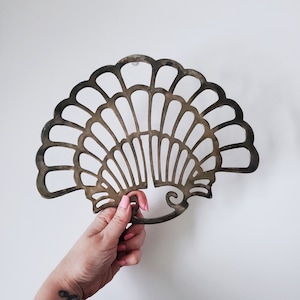 May include: A decorative metal trivet in the shape of a fan with a scalloped design. The trivet is made of a light brown metal and has a rustic, vintage look.