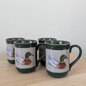 May include: Four ceramic mugs with a green and gold trim. Each mug displays a mallard duck illustration on a light grey panel. The mugs have a dark green handle and base. Ideal for tea or coffee, these mugs are a charming addition.