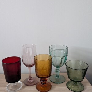 May include: A collection of five colorful drinking glasses. The glasses are in shades of red, pink, amber, teal, and green. They vary in shape, including stemmed wine glasses and tumblers. The glasses are arranged on a light-colored wooden surface.