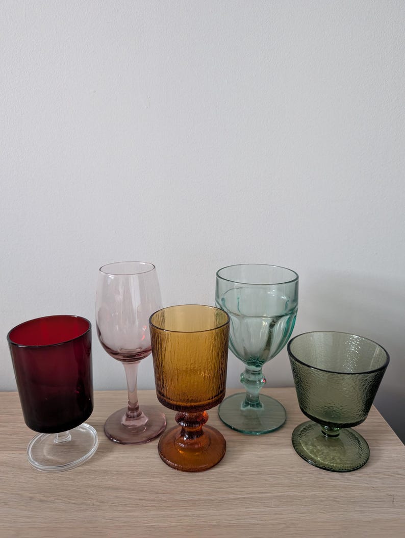 May include: A collection of five colorful drinking glasses. The glasses are in shades of red, pink, amber, teal, and green. They vary in shape, including stemmed wine glasses and tumblers. The glasses are arranged on a light-colored wooden surface.