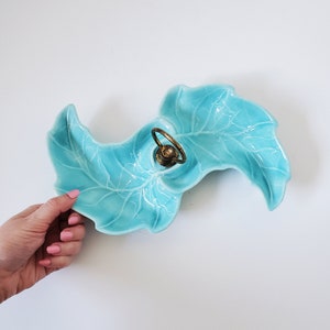 May include: A turquoise ceramic serving dish shaped like two leaves with a gold metal ring in the centre.
