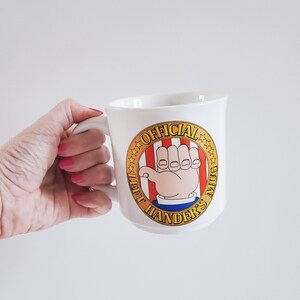 May include: White ceramic mug with a large handle, featuring a cartoon hand with the text "OFFICIAL LEFT HANDER'S MUG" in a circular design with red and white stripes and gold stars.