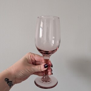 May include: A pink-tinted wine glass with a rounded bowl and a long stem. The glass is held up against a plain background. The glass is made of clear glass and has a delicate appearance. The stem and base are a darker shade of pink.