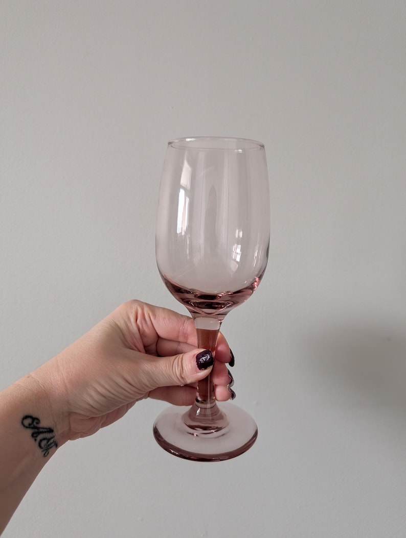 May include: A pink-tinted wine glass with a rounded bowl and a long stem. The glass is held up against a plain background. The glass is made of clear glass and has a delicate appearance. The stem and base are a darker shade of pink.