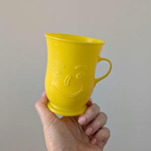May include: A bright yellow mug with a smiling face design. The mug has a handle and a rounded base. The face features curved eyebrows, a nose, and a wide smile. The mug is held in a hand.