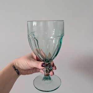 May include: A clear, teal-colored goblet with a ribbed design on the bowl and a circular base. The stem is short and the glass has a vintage aesthetic. The glass is held by a person.