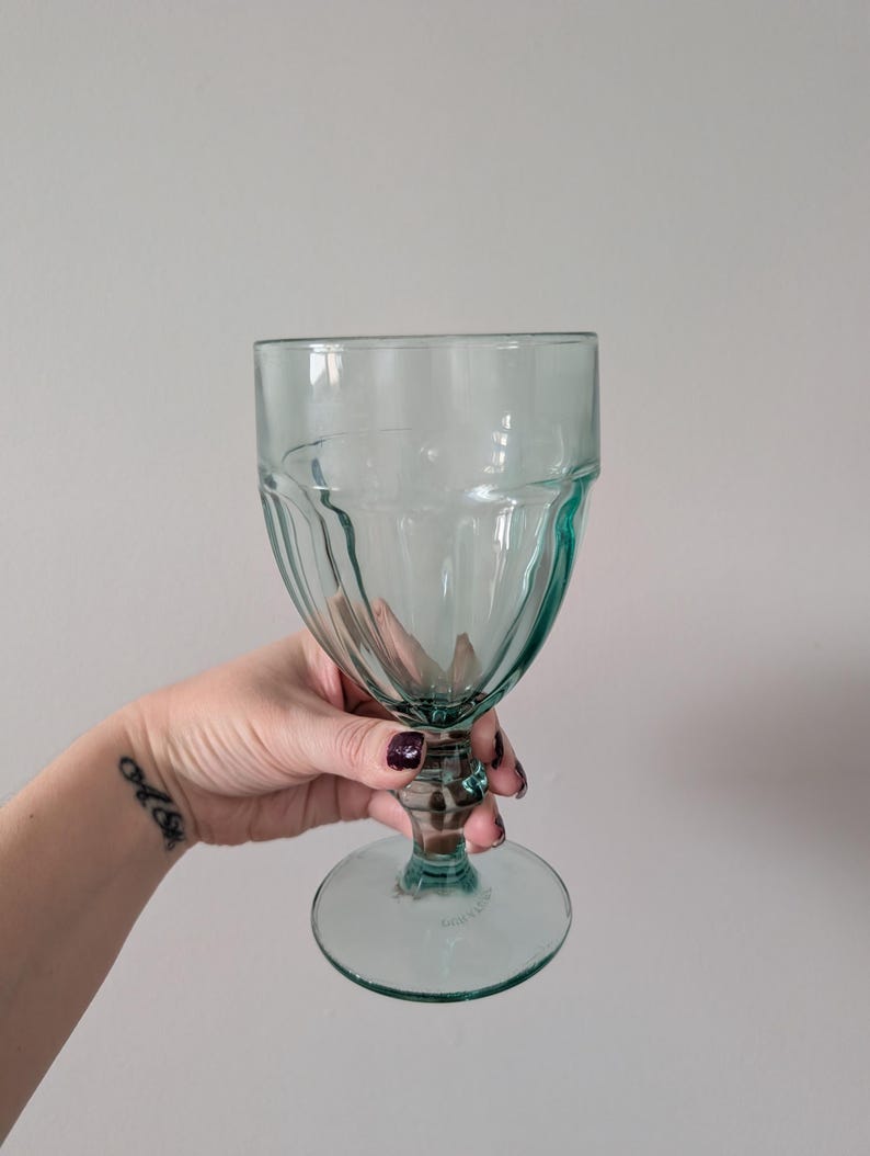 May include: A clear, teal-colored goblet with a ribbed design on the bowl and a circular base. The stem is short and the glass has a vintage aesthetic. The glass is held by a person.