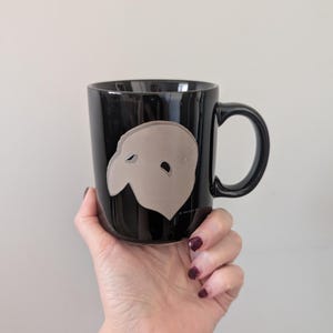 May include: Black ceramic mug featuring a light gray mask design. The mug has a black handle and a glossy finish. The design is a stylized representation of a theatrical mask. The mug is held by a person.