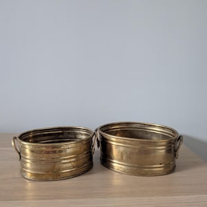 Puede incluir: Dos jardineras ovaladas antiguas de latón con asas pequeñas. Las jardineras tienen un diseño acanalado y un color dorado cálido. Se colocan una al lado de la otra sobre una superficie de madera clara, lo que sugiere un uso decorativo o funcional.