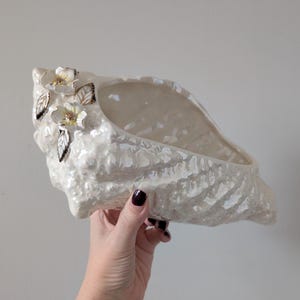 May include: A pearlescent white ceramic seashell planter with a textured surface. The planter is adorned with white flowers and gold-colored accents. The seashell is held by a person.