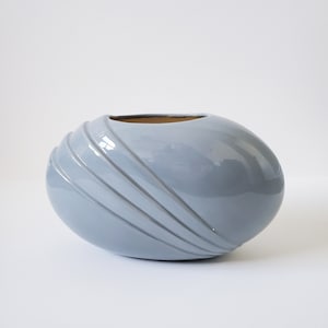 May include: A light blue ceramic vase with a rounded oval shape and a smooth, glossy finish. The vase has a series of raised, horizontal lines on the front.