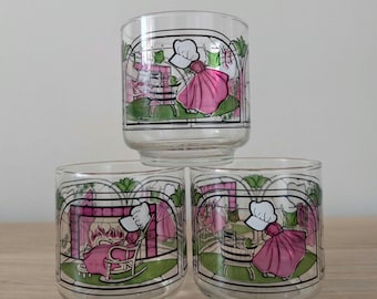 May include: Three clear glass tumblers featuring colorful screen-printed illustrations. The designs depict figures in pink dresses and white bonnets engaged in domestic activities like rocking in a chair by a fireplace and washing laundry.