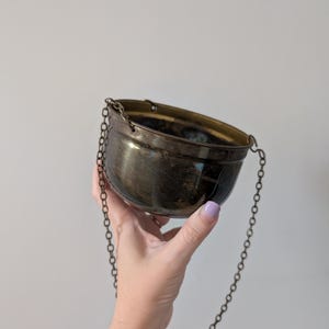May include: A small, round, metallic bowl with a dark, polished finish and a reflective surface. The bowl features two small loops on opposite sides, from which a thin, delicate metal chain is attached, allowing it to be suspended or hung.