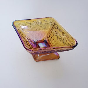 May include: A square, iridescent glass candy dish with a pedestal base. The dish has a floral pattern and is a vibrant amber color.