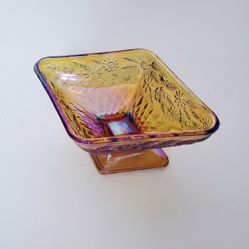 May include: A square, iridescent glass candy dish with a pedestal base. The dish has a floral pattern and is a vibrant amber color.