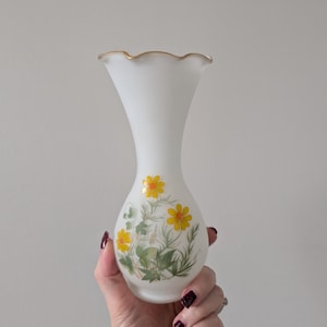 May include: White glass vase with a ruffled gold rim. The vase features a floral design with yellow flowers and green leaves. The vase is held in a hand against a plain background.