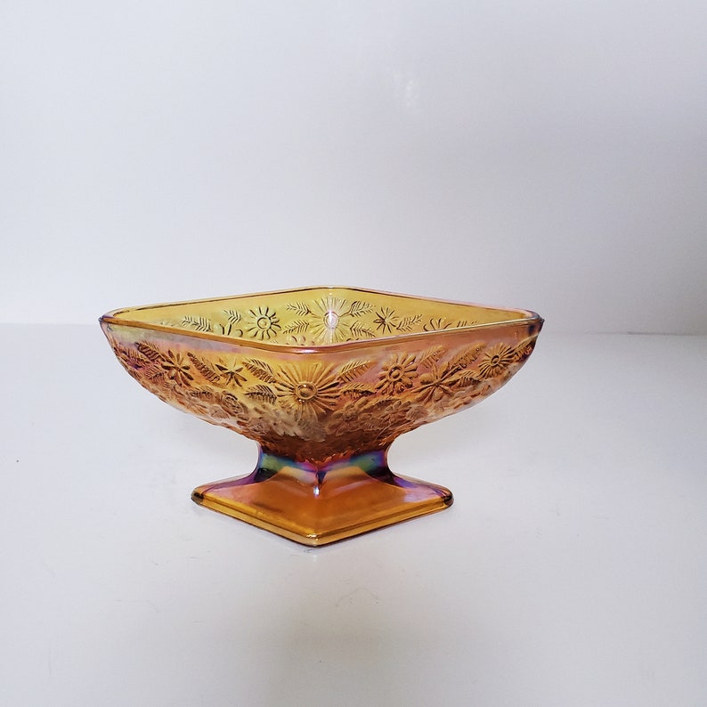 May include: A square, amber-colored glass bowl with a pedestal base. The bowl has a floral design and iridescent finish.