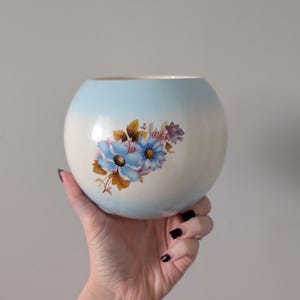 May include: A spherical ceramic vase with a light blue and white gradient. The vase has a floral design with blue and purple flowers. The vase is held against a plain, light grey background.