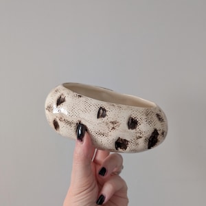 May include: Cream-colored ceramic bowl with a speckled pattern of brown spots. The oval-shaped bowl has a textured surface and a smooth, off-white interior. A decorative piece for the home.