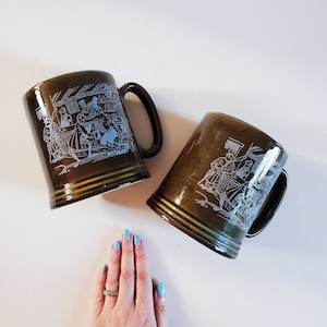 May include: Two brown ceramic mugs with white illustrations of people in a tavern scene. The mugs have a dark brown glaze and a gold rim.