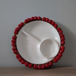 Könnte beinhalten: Weißes Keramik-Serviertablett mit drei Fächern und einer kleinen runden Dip-Schale. Der Rand ist mit einem Kranz aus roten, beerenartigen Verzierungen verziert. Das Tablett steht auf einer Holzoberfläche.