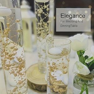 May include: Glass vases filled with water and gold beaded decorations. The vases contain floating candles and white roses, with the text "Elegance For Wedding And Dinning Table".