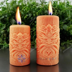 Può includere: Due candele a colonna arancioni accese con intricati disegni intagliati. Le candele cilindriche presentano un motivo a rilievo dettagliato. Le fiamme sono luminose e lo sfondo include un fogliame verde. Le candele sono di diverse dimensioni.