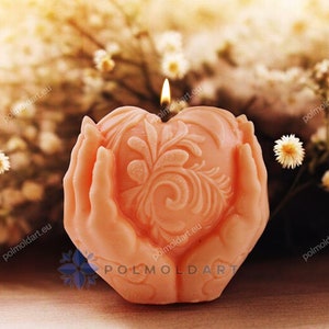 May include: A peach-coloured candle in the shape of a heart, held by two hands. The heart is decorated with an ornate floral design. A small flame burns atop the candle, and white flowers are in the background.
