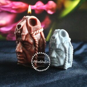 May include: Two sculpted candles in the shape of a Native American chief, one lit with a flame. The larger candle is brown, the smaller one is gray. Both have a detailed headdress and facial features. The background is blurred with pink and green hues.