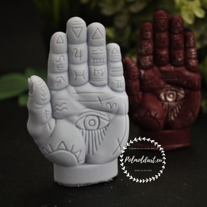 May include: Two hand-shaped sculptures, one light gray and one maroon, are displayed. The gray sculpture features an eye design and various symbols. The maroon sculpture has an eye design. The background is dark with greenery.