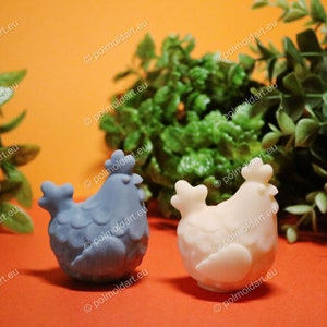Può includere: Due statuette a forma di gallina, una azzurra e una bianco sporco, sono su una superficie arancione. Le galline sono decorative, con dettagliate fantasie di piume. Sullo sfondo c'è del fogliame verde. Il sito web "polmoldart.eu" è in filigrana.