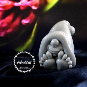 May include: A gray gnome figurine with a pointed hat and large feet. The gnome is smiling and appears to be hiding under its hat. The figurine is made of a smooth material and is set against a dark blue background.