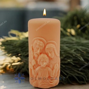 Puede incluir: Una vela de pilar encendida de color melocotón con un relieve tallado que representa una escena familiar religiosa. La vela está colocada sobre una superficie de madera con follaje verde en el fondo. La llama de la vela es visible.