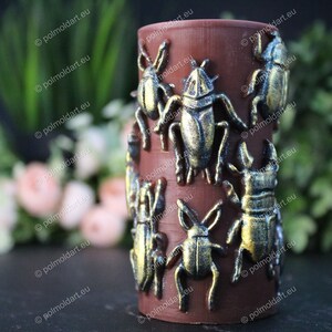 Pu&ograve; includere: Un portacandele cilindrico marrone adornato con decorazioni di scarabei color bronzo. Gli insetti sono disposti attorno all'esterno del portacandele. Lo sfondo presenta vegetazione e fiori sfocati.