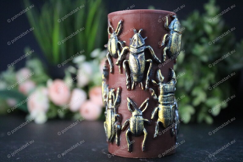 Pu&ograve; includere: Un portacandele cilindrico marrone adornato con decorazioni di scarabei color bronzo. Gli insetti sono disposti attorno all'esterno del portacandele. Lo sfondo presenta vegetazione e fiori sfocati.