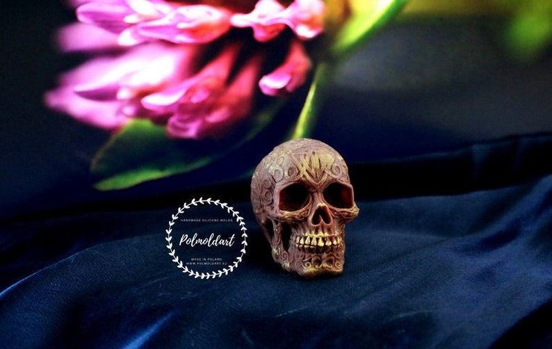 May include: A detailed, brown and gold skull figurine with intricate carvings. The skull is set against a dark blue background with a blurred pink and green floral element. The Polmoldart logo is visible.