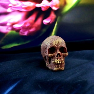 May include: A detailed, brown skull figurine with intricate carvings. The skull has open eye sockets and a detailed jawline. The background features a blurred pink flower and a dark blue fabric.