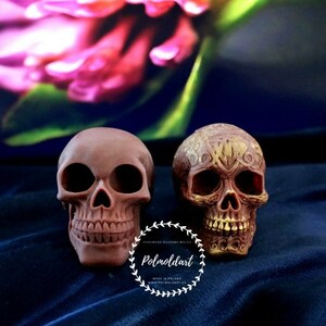 May include: Two brown skull figurines. One is plain, the other is decorated with an ornate gold design. The skulls are set against a dark blue background with a blurred floral element. The text "Polmoldart" is visible.